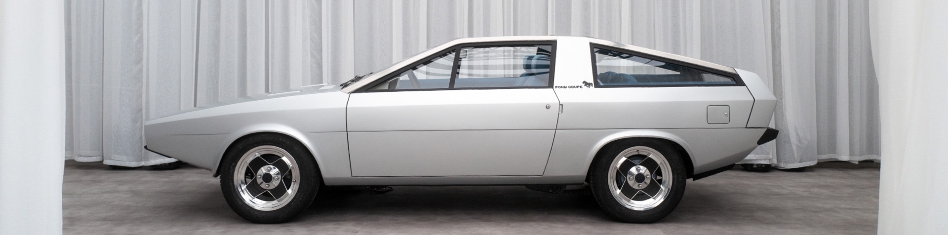 Side view of the restoration of the Pony Coupe Concept car in grey with silver hubcaps presented on a stage with white curtains.