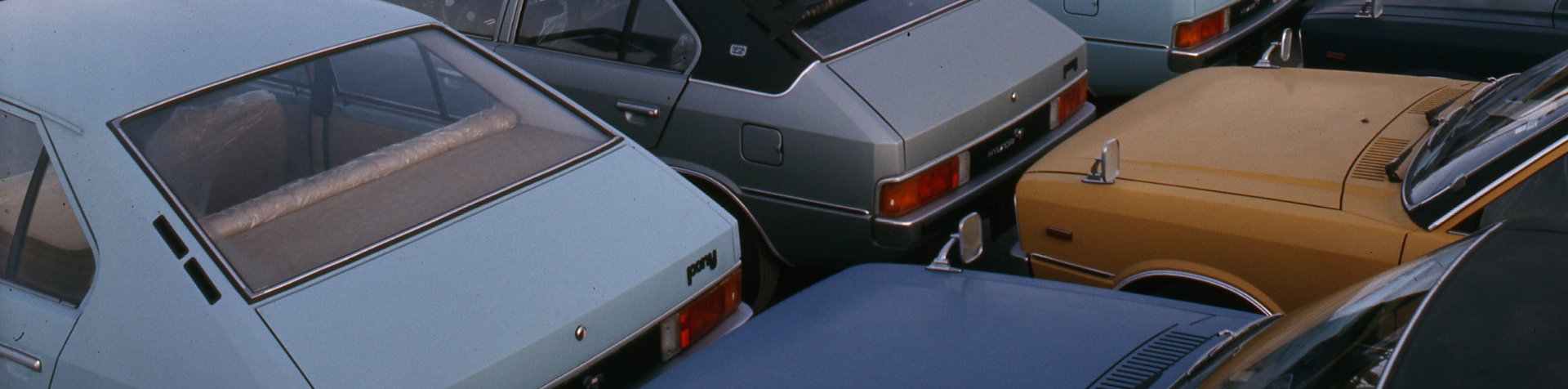 Closeup of six colorful early-model Hyundai Ponys parked tightly and neatly in two rows at a car terminal.