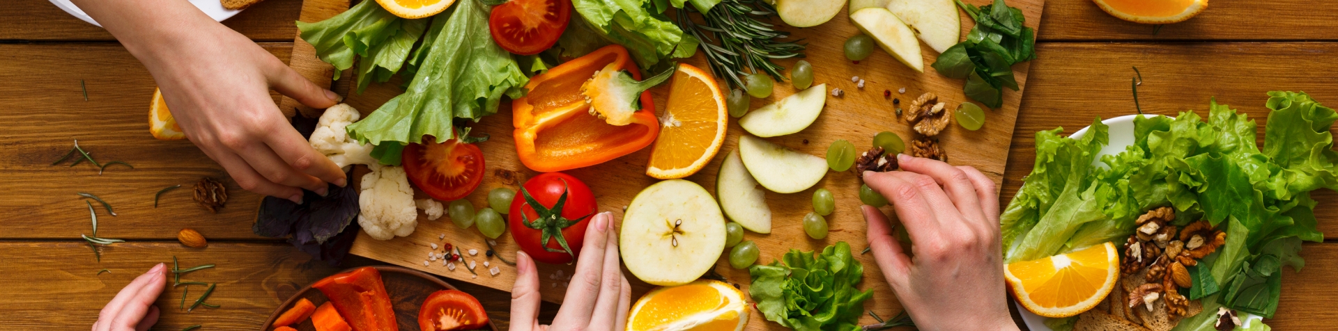 A large wooden chopping board covered in chopped-up fruits, vegetables, and herbs. There are three hands from unseen people arranging the food including grapes, apples, oranges, lettuce, cherry tomatoes, and rosemary.