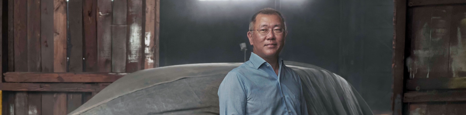 Hyundai Motor Group Executive Chair Euisun Chung standing in a warehouse under a spotlight in front of a car covered in a sheet. He is wearing a blue collared shirt and oval-shaped glasses.