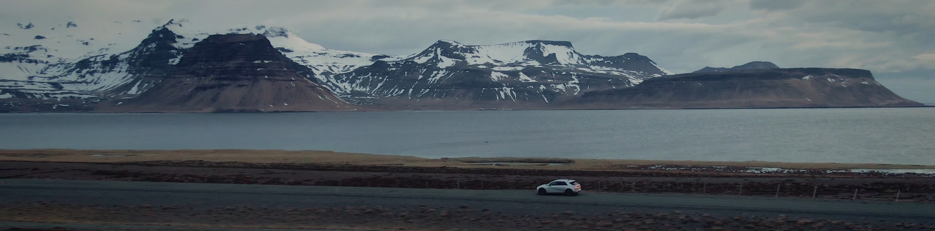 A gray IONIQ 5 drives along a road beside a lake with snow-capped mountains in the background.