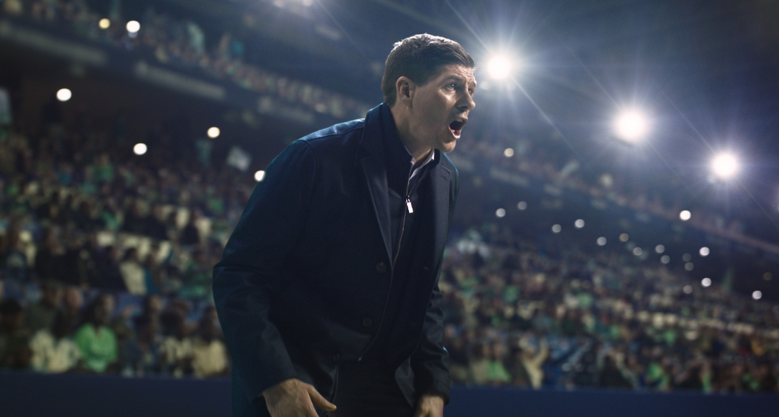 Steven Gerrard dressed in a dark coat coaching his team from the side lines inside a stadium.