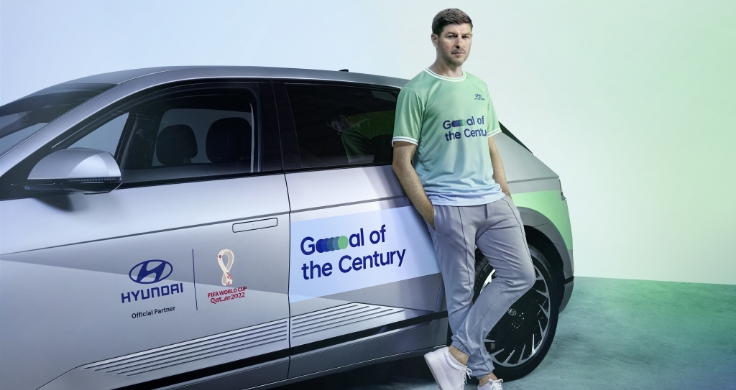 Three-quarter view of a silver Hyundai IONIQ 5 with “Hyundai” and “FIFA World Cup 2022” stickers on the front door and a “Goal of the Century” sticker on the rear door next to which Steven Gerrard is leaning against the car wearing a blue and green Goal of the Century shirt.