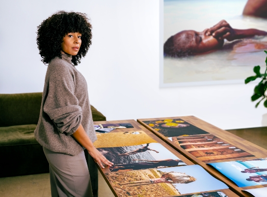 Nicky Woo surveying her photographs and looking behind her. The photographs are laid out on the table in front of her, she is wearing brown pull-over and brown trousers and behind her there is a green couch.